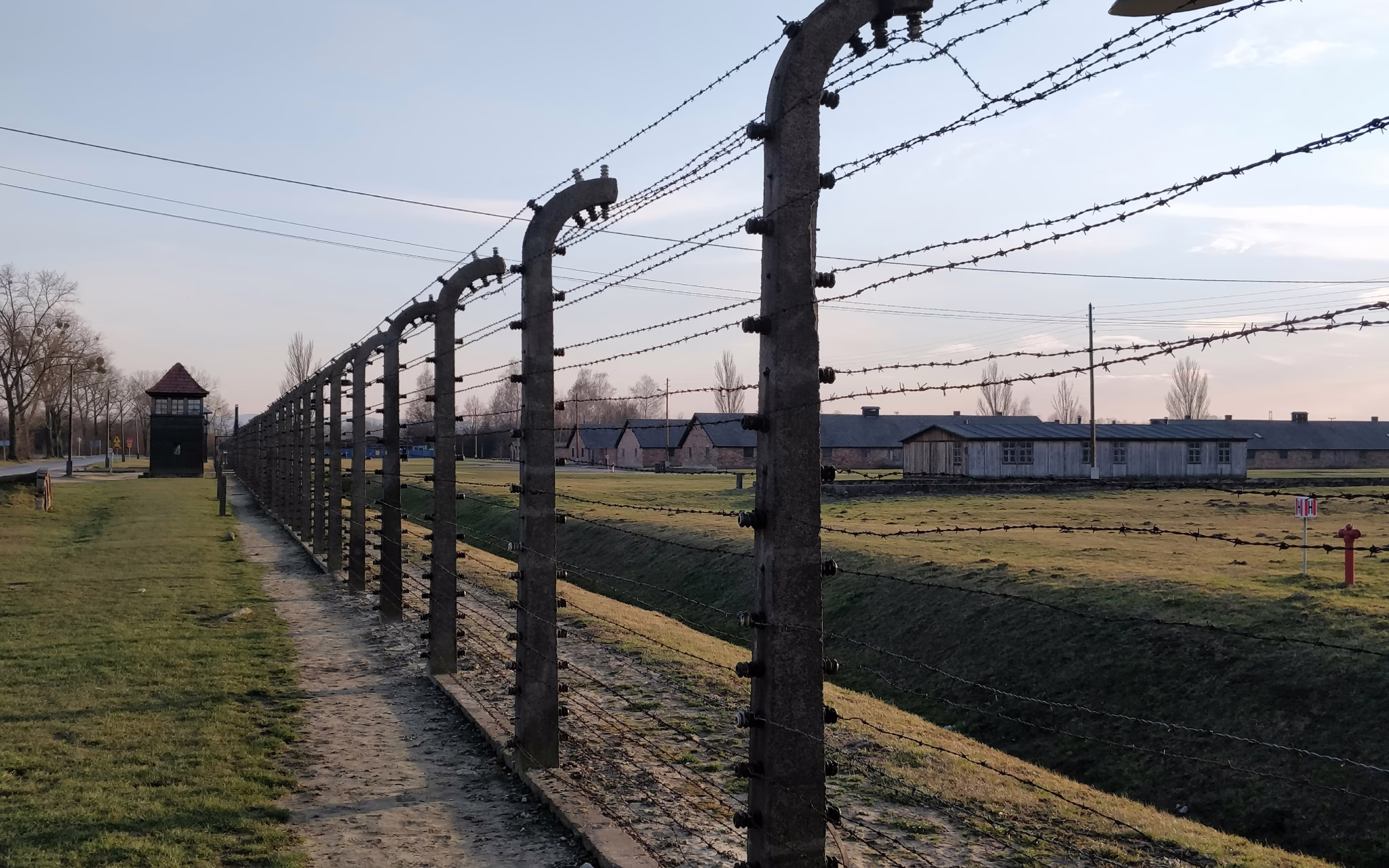 Bild eines Zauns im KZ Auschwitz (Oświęcim, Polen) mit einzelnen Häusern im Hintergrund